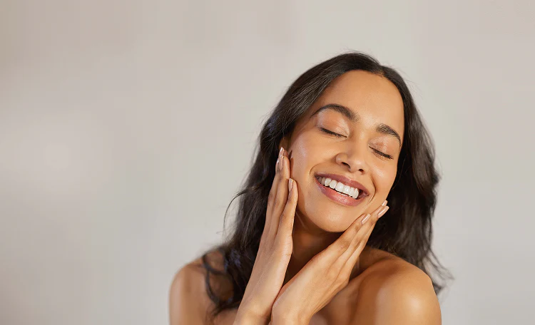 Mujer sonriendo con los ojos cerrados, tocándose suavemente las mejillas con las manos